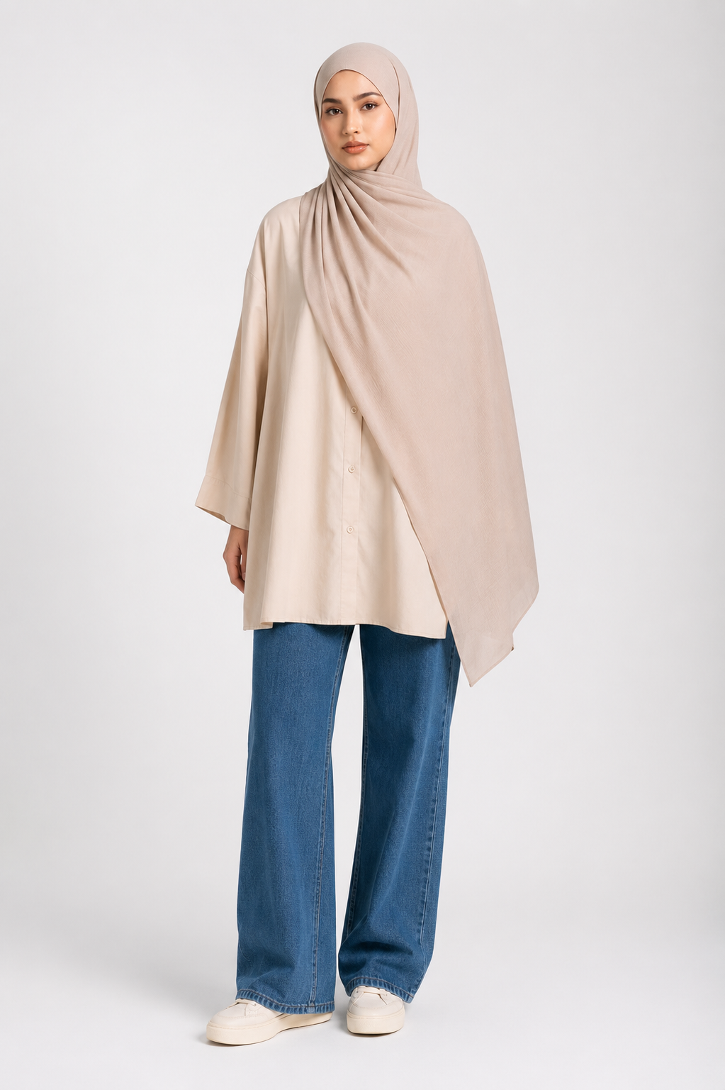 A woman modeling a light beige modal hijab set draped over a matching beige tunic, paired with blue jeans and white sneakers, against a light gray studio background.