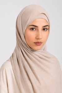 A woman wearing a draped light beige modal hijab and matching top, photographed against a plain light gray studio background.