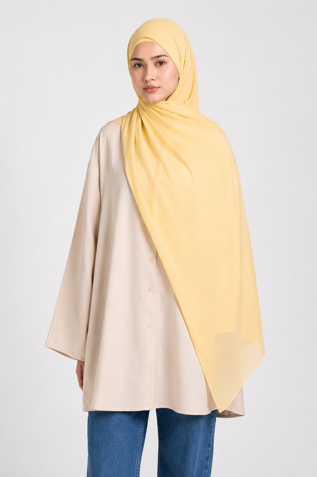 A woman wearing a light yellow modal hijab and a beige button-up tunic with blue jeans, photographed against a plain light gray studio background.